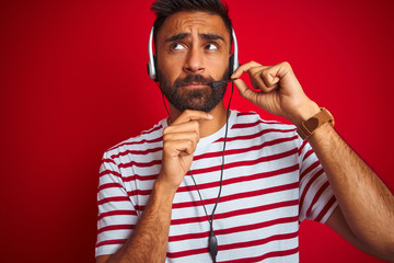 Wall Mural - Young indian call center agent man using headset over isolated red background serious face thinking about question, very confused idea