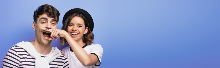 panoramic shot of cheerful girl holding finger with drawn mustache near face of man on blue background