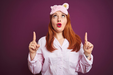 Sticker - Young beautiful redhead woman wearing pajama and mask over purple isolated background amazed and surprised looking up and pointing with fingers and raised arms.