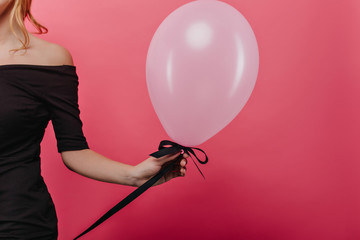 Studio shot of slim white woman holding helium pink balloon. Party photo of graceful female model in black dress posing on bright background.