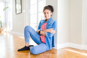 Beautiful young african american woman with afro hair sitting on the floor happy face smiling with crossed arms looking at the camera. Positive person.