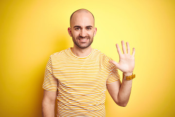 Young bald man with beard wearing casual striped t-shirt over yellow isolated background showing and pointing up with fingers number five while smiling confident and happy.