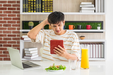 A young Asian boy siting with laptop holding a tablet with orange juice and a plate of vegetable snack frustrating expression.