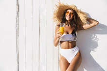 Photo Of Caucasian Brunette Woman In Straw Hat And Sunglasses Smiling While Standing Over White Wall With Orange Juice