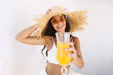 Poster - Photo of cheerful brunette woman in straw hat and swimsuit smiling while standing over white wall with orange juice