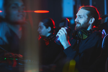 Wall Mural - Smiling satisfied young bearded gamer with headphones on neck sitting in comfortable armchair and drinking energy beverage while playing video game