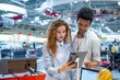 © pantovich - Red-haired girl with glasses with an African guy in an electronics store standing in front of the stand choosing a tablet computer to buy