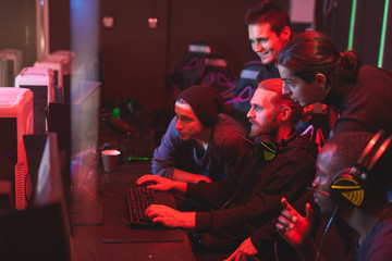 Wall Mural - Group of focused young men standing together in front of computer monitor and playing video game, friends assisting guy to pass network game