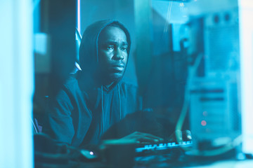 Wall Mural - Serious tired black man in hoodie sitting in dark blue room and typing on computer keyboard while working on system software