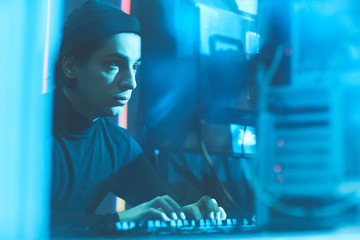 Wall Mural - Serious concentrated young Jewish hacker in beanie hat sitting at table in dark room and typing on keyboard while breaking into server data