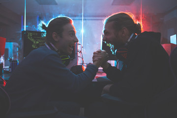 Cheerful young hipster software engineers with headsets on necks sitting in database center and shaking hands while being excited