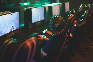 Row of serious concentrated guys in headsets sitting in gaming chairs and playing net game