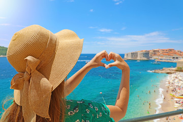 Young beautiful woman in Dubrovnik making hands shaped heart on spring summer warm sunny day. Girl with hat happy outdoors in her travel in Southern Croatia, Europe.