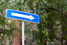 Right Direction Allowed One Way Blue Street Sign On A City Street, Against The Gray Buildings And Cloudy Sky Bokeh Background