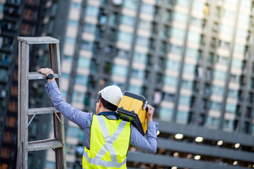 Asian maintenance worker man with safety helmet and green vest carrying aluminium step ladder and tool box at construction site. Civil engineering, Architecture builder and building service concepts