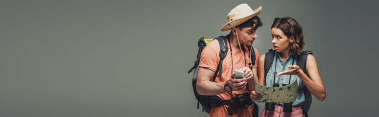 panoramic shot of two young tourists talking while looking at geographic map on grey background