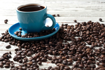 Full blue ceramic cup of black hot coffee near lot of scattered fried beans on old rustic white wooden table. Copy space for text