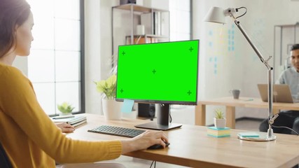Over the Shoulder: Creative Young Woman Sitting at Her Desk Using Desktop Computer with Mock-up Green Screen. Bright Office where Diverse Team of Young Professionals Work on Computers