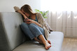 Portrait of depressed young woman hiding her face sitting on grey textile couch holding the phone. Cyber bullying victim concept. Sad female in her room. Background, copy space.