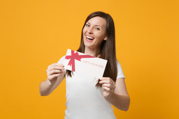 Portrait of cheerful attractive young woman in white casual clothes holding gift certificate isolated on bright yellow orange wall background in studio. People lifestyle concept. Mock up copy space.