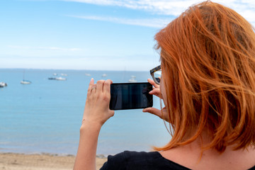 Beautiful young red hairs woman making photo with smartphone on beach