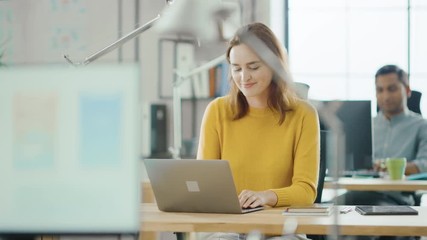Beautiful and Creative Young Woman Sitting at Her Desk Using Laptop Computer. In the Background Bright Office where Diverse Team of Young Professionals Work 