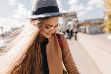 Wall Mural - Romantic fair-haired girl in beige outfit looking down with smile. Outdoor photo of graceful woman in gray hat decorated with black ribbon enjoying sunny day in autumn.