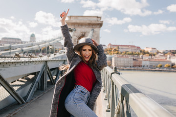 Wall Mural - shapely blissful girl in red sweater dancing on bridge on blur city background in autumn morning. ou