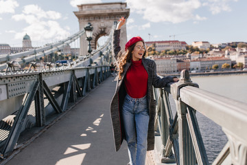 Wall Mural - joyful french female traveler in red beret looking at river from bridge. outdoor portrait of relaxed