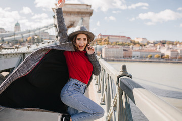 Wall Mural - excited long-haired woman dancing in long coat and laughing on urban background. slim girl in red sw
