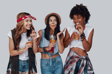 Poster - Three attractive young women drinking refreshing cocktail and smiling while standing against grey background