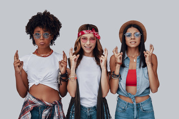 Poster - Three attractive young women keeping fingers crossed and looking at camera while standing against grey background