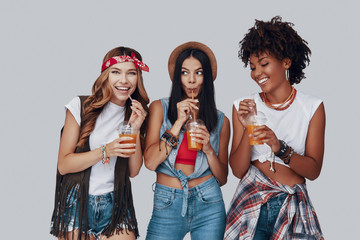 Canvas Print - Three attractive young women drinking refreshing cocktail and smiling while standing against grey background