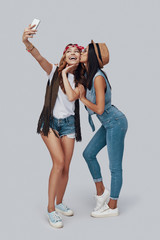 Canvas Print - Full length of two attractive stylish young women taking selfie and smiling while standing against grey background