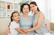 © DragonImages - Portrait of happy Asian family generation mother daughter and grandmother embracing and smiling at camera while resting on couch