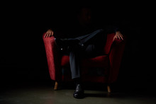 Male Businessman In A Black Suit Sitting In Red Chair, Black Background, No Faces Visible, Studio Shooting