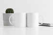Two mugs mockup with a notebook and a cactus in a pot on white table.