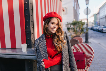 Wall Mural - Beautiful white woman in soft coat posing near cafe with striped exterior. Outdoor portrait of pretty caucasian girl wears beret standing with cup of coffee and looking with interested smile.