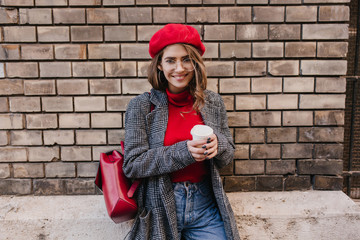 Wall Mural - Amazing french lady in tweed coat drinks latte on brick wall background. Sensual stylish woman in red beret posing with little leather backpack in autumn day.