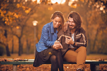 Wall Mural - Young couple using cellphone in autumn colored park.