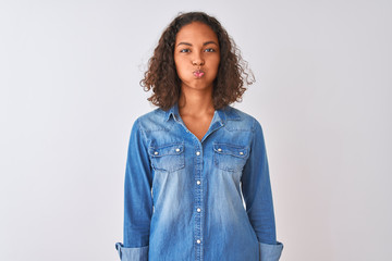 Canvas Print - Young brazilian woman wearing denim shirt standing over isolated white background puffing cheeks with funny face. Mouth inflated with air, crazy expression.