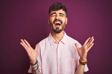 Young man with tattoo wearing summer pink shirt standing over isolated purple background celebrating mad and crazy for success with arms raised and closed eyes screaming excited. Winner concept