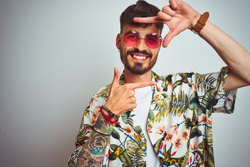 Man with tattoo on vacation wearing summer shirt sunglasses over isolated white background smiling making frame with hands and fingers with happy face. Creativity and photography concept.