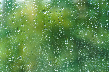 Close up of fresh water drops on window glass on background of green leaves. Raindrops on windowpane in summer day. Droplets on glass with blurry summer garden background.