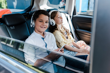 Wall Mural - selective focus of boy using smartphone and listening music in headphones near sister in car