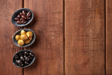 Various Types Of Olives In Bowls, Shot From The Top On A Dark Rustic Wooden Background With Copy Space. Purple, Green Almond Stuffed, And Black Pitted Olives With A Place For Text