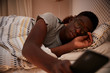 Millennial African American man half asleep in bed, wearing glasses and holding smartphone, close up