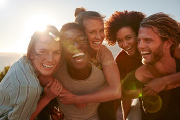 Five millennial friends on a road trip have fun piggybacking at the roadside, front view, lens flare