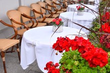 Empty Street Cafe With White Tablecloth And Bouquet Of Red Flowers On The Table, Row Of Cozy Chairs On Background And Blurred Red Geranium Flower On The Front. Beautiful Restaurant In The Old Town