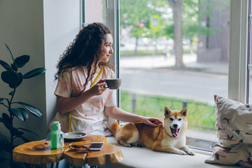 Happy student pretty girl is stroking adorable shiba inu dog sitting in cafe on window sill with cup of tea and enjoying leisure time. People and relaxation concept.